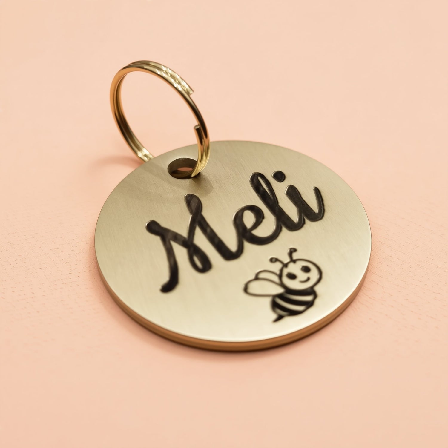 A round metal keychain with the name "Meli" engraved on it, featuring a small bee illustration.